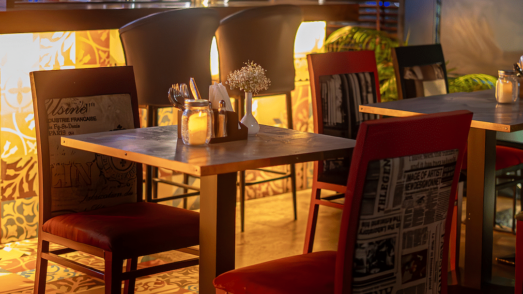 A warmly lit restaurant interior with a bar in the background and tables and chairs in the foreground. The bar has a white counter and hanging lightbulbs. The tables have candles and flowers on them, and the chairs are upholstered in red fabric with newspa